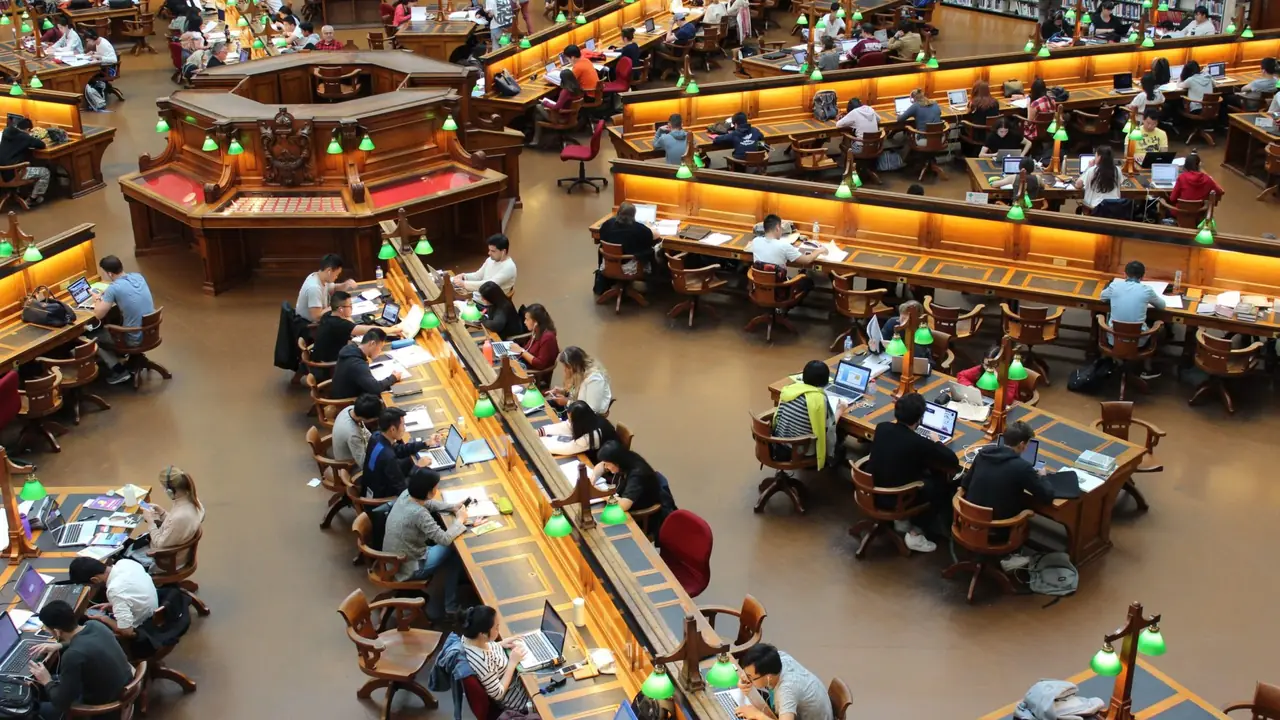 Students working at long shared desks in a busy tiered university library