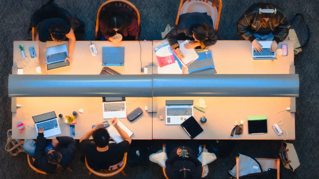 Overhead view of seven students studying at two long library desks covered with laptops, notebooks and phones.