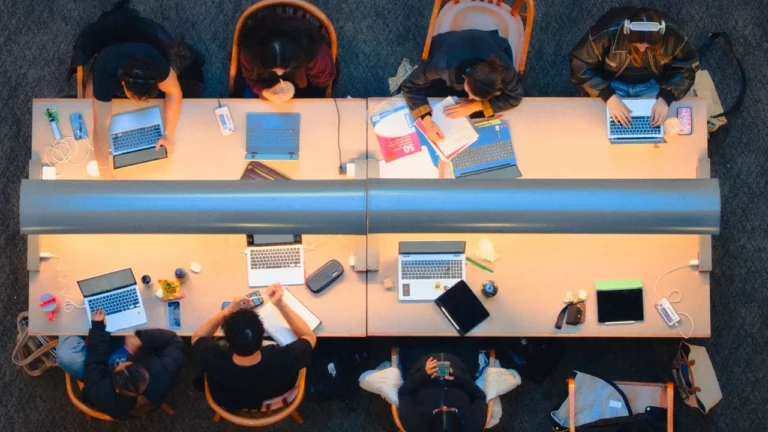 Overhead view of seven students studying at two long library desks covered with laptops, notebooks and phones.