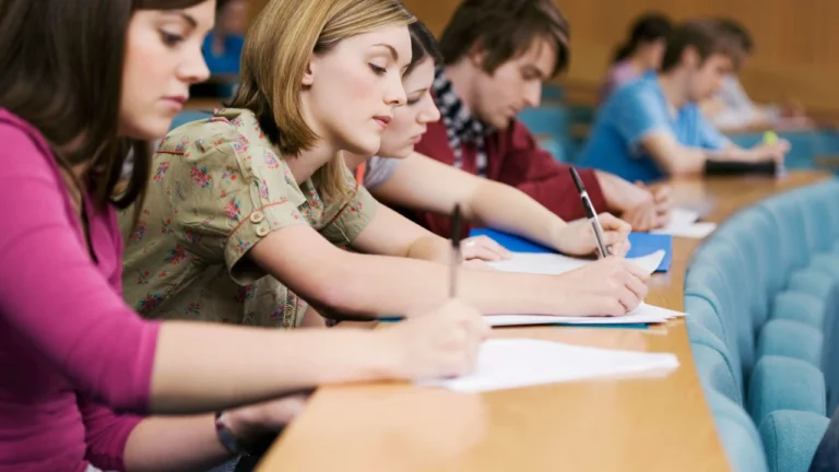 Students sitting in a row at a long desk writing in notebooks during a class