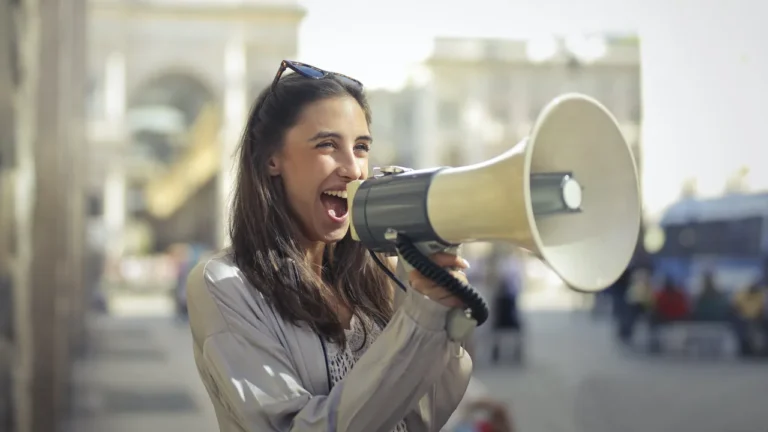 A smiling young woman with sunglasses pushed up on her head shouts into a large white megaphone on a sunny city street, with blurred stone buildings and passers-by behind her.