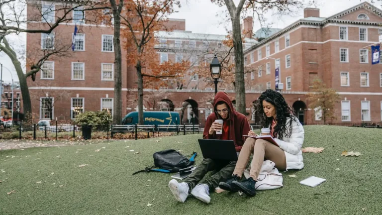 Two students sitting on a lawn with a laptop in front of a red brick campus building