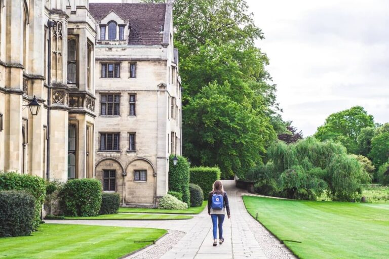 A student walking with a backpack along a path beside a large stone collegiate building with mullioned windows, lawns and mature trees.