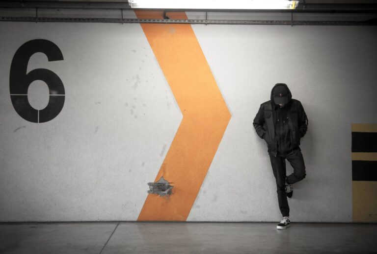 A person in a black hooded jacket and cap leaning against a car-park wall, beside a large painted orange chevron and the numeral 6.