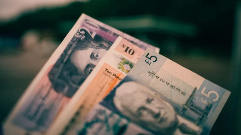 Close-up of British banknotes fanned out together: a 20 pound, a 10 pound and a 5 pound note, against a blurred dark green background.