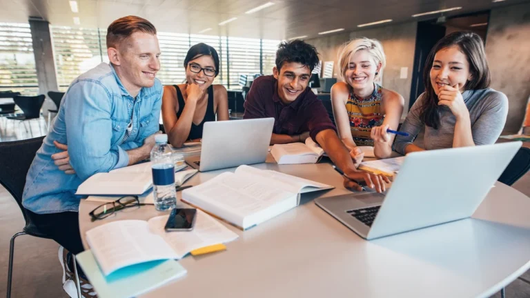 Five students gathered around a table in a study room, smiling at a laptop screen, with open textbooks and notebooks spread out in front of them.