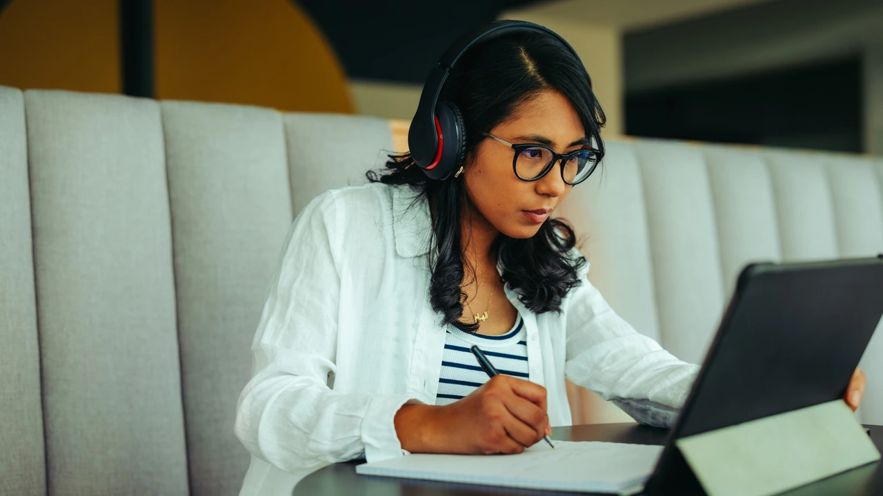 A student wearing over-ear headphones and glasses writing in a notebook while looking at a tablet propped up on a cafe table.