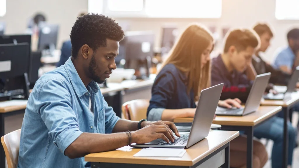 A student in a denim shirt typing on a laptop at a desk in a computer lab, with three other students working at laptops in the row behind him.