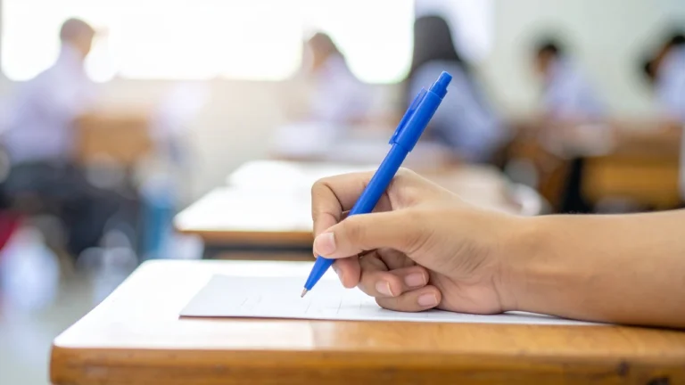 A close-up of a student's hand writing on an answer sheet with a blue ballpoint pen at a wooden desk, with other candidates blurred in the rows behind.
