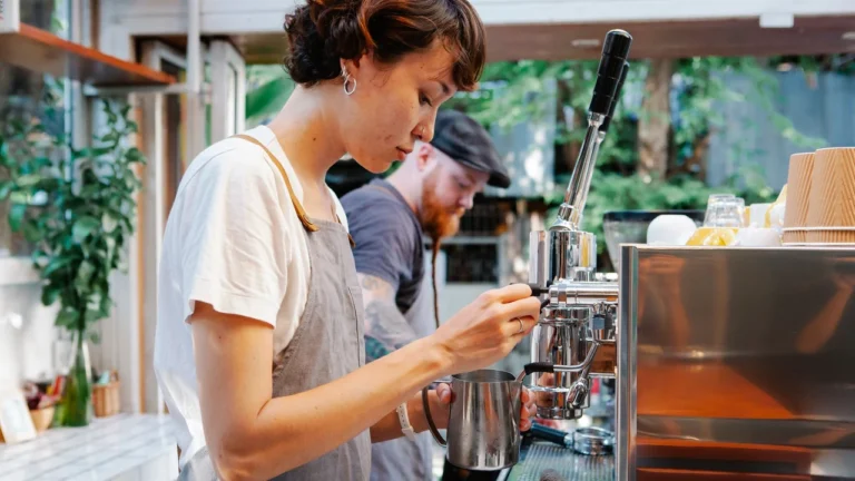 A young barista in a white T-shirt and grey apron steams milk at a chrome espresso machine, while a second barista works behind her in a bright cafe with green plants.