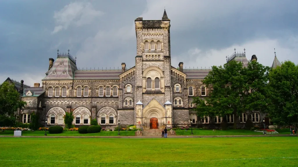 The Gothic Revival stone front of University College at the University of Toronto, with its central tower and arched wooden doorway, seen across a wide green lawn under a grey, cloudy sky.