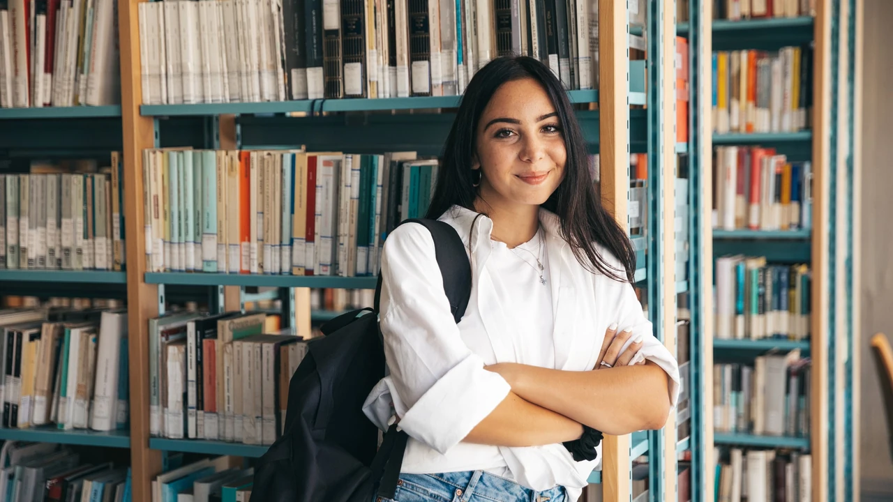 A student with a backpack over one shoulder standing with arms folded in front of tall library shelves packed with books.