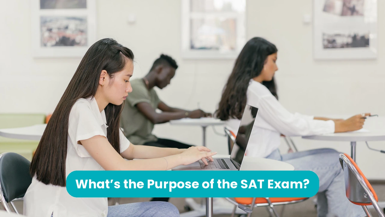 A student with long dark hair types on a laptop at a round white table in a bright classroom, while two other students work at desks behind her. A teal banner across the lower third reads ‘What’s the Purpose of the SAT Exam?’.